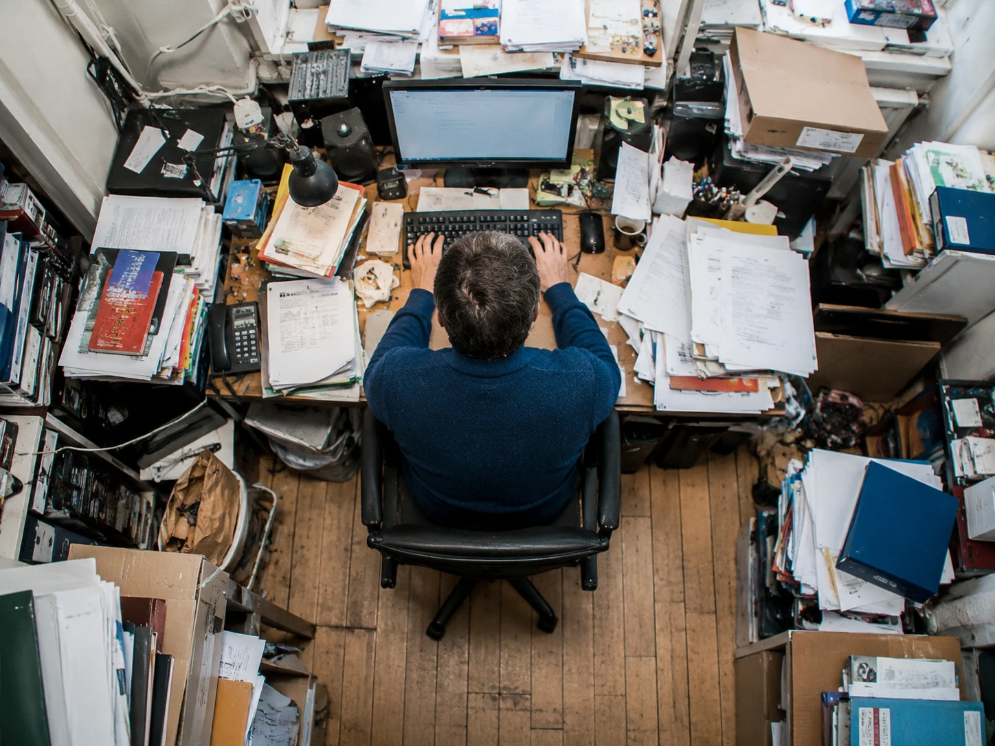 Person sitting at the centre of a cluttered office desk, viewed from above, surrounded by documents, folders, books and office supplies.